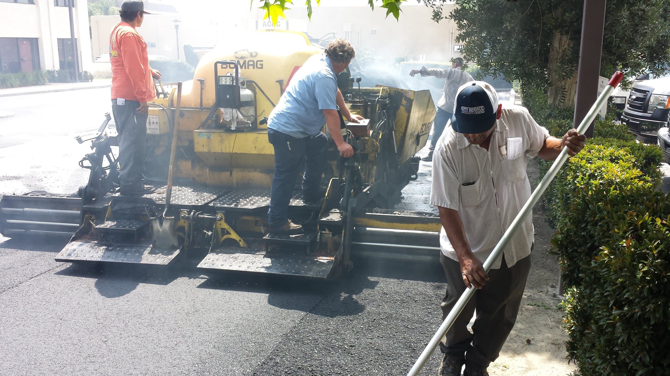 Paving crew operating an asphalt paver while a worker rakes the fresh mat by hand